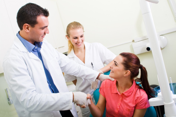 A male dentist and his female assistant shake the hand of a female patient inside a dental office